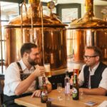 Schwangau Schlossbrauhaus: Zwei Männer sitzen mit Tracht in einem Brauhaus mit Braukesseln im Hintergrund